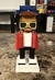 A blocky, stylized figure made of plastic, resembling a person with sunglasses, a blue hat, a red jacket, and black pants. The figure stands on a white base, and the setting is indoors with a marble-like floor surface and furniture visible in the background.