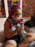 A woman with bright pink braids sitting on a bed and holding a bouquet of colorful flowers close to her face. She is wearing a white tank top and dark shorts, smiling softly at the camera. The background shows a brick wall and part of a bed frame.