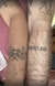 A close-up of two forearms placed side by side, both featuring tattoos. The left arm has a tattoo an ash tree branch. The right arm has the word "WOODLAND" tattooed in capital letters.