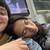 A person is resting their head on their partner's shoulder inside a public transit vehicle. The resting person has long dark hair and their eyes closed, while the other person is partially visible, smiling.