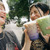 A young couple laughing and holding up drinks in clear cups with straws. They are standing outdoors on a street with trees and cars in the background, sharing a joyful moment together.