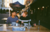 A young couple sitting outdoors at a wooden table, sharing a meal. The man on the left is wearing a blue sweater and glasses, holding chopsticks, while the man on the right is wearing a black sweater with a white stripe. They are smiling at each other, with a street view in the background.