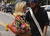 A young couple standing close together on a city street. The woman on the left has blonde braids and is holding a bouquet of flowers. The man on the right is wearing a bandana and a black shirt, looking affectionately at his partner.