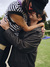 A young couple embracing closely. The person in the striped shirt is being lifted off the ground by their partner who is wearing a leather jacket. They are outdoors, surrounded by green grass and some flowers in the background.