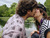 A young couple sharing a kiss near a tranquil pond surrounded by greenery. The person on the left is wearing a plaid shirt, and the person on the right is wearing a black hat. Ducks can be seen swimming in the pond.