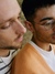 A close-up shot of a young couple sitting closely together, sharing a quiet moment. The man on the left has a light beard and is wearing a white sweater with black stripes, and the man on the right is wearing glasses and an orange sweater over a white shirt.