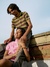 A young souple are sitting on a low brick wall, smiling and laughing. The person standing behind is wearing a green and purple striped polo shirt and gray jeans, leaning over the person in front, who is wearing a pink jacket and holding onto the legs of the person behind them. The sky is clear and blue in the background.