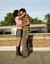 A young couple are standing close together on a rooftop, embracing and about to kiss. The person on the left is wearing a pink jacket, gray shorts, and brown boots, while the person on the right is wearing a green and purple striped shirt with gray jeans. A train is visible in motion in the background, along with green trees and a clear sky.