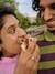 A close-up shot of a young couple smiling warmly at each other while sharing a slice of pizza. The person on the left is in the process of taking a bite, with a playful and happy expression. They are wearing a pink top, and their focus is on the pizza. The person on the right is looking at them with a wide smile, wearing a green and purple striped shirt. The background is slightly blurred, showing greenery and a bit of the sky.