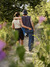 A young couple walking down a sunlit path surrounded by greenery. They have their arms around each other, one person’s hand resting on the other’s hip. The scene is framed by wildflowers, giving it a serene and romantic atmosphere