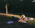 A young couple are lying on a blanket in a park, surrounded by grass and trees. One person is lying on their back, while the other person is sitting up, leaning over them, and gently touching their face. The scene is lit by soft, warm sunlight, creating a peaceful and intimate atmosphere.