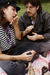 A couple having a picnic on the grass. The person on the left is licking their fingers while holding a piece of bread with jam. The person on the right, wearing a leather jacket and glasses, watches them with a smile. The picnic setting includes a blanket and jars of jam.