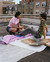 A young couple is sitting on a rooftop, casually enjoying pizza. The person on the left is crouching and smiling, holding a slice of pizza. They are wearing a pink top, dark shorts, and brown boots. The person on the right is reclining with their legs stretched out, wearing a green and purple striped shirt and grey pants. They are sitting on a white and pink blanket spread out on the rooftop, with two open pizza boxes in front of them, containing partially eaten pizzas. The background shows a row of brick buildings.