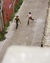 A young couple are running down a deserted alleyway with red brick walls and large red doors on the left side. The person on the left is wearing a green striped shirt and gray pants, while the person on the right is wearing a pink shirt and black shorts. The alley is lined with greenery and graffiti is visible on the walls.