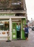 A young couple are standing outside a sweet shop with a green door. The man is in a turquoise sweater and is holding a box of sweets, about to eat one. The woman, with red hair and a furry hat, is smiling. The shop is located on Brick Lane, with the street sign visible above.