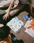 An overhead view of a chess game in progress on a checkered board with blue and white pieces. A couple are sitting on the ground next to the board, one wearing beige pants and the other in an orange sweater. There are also books, newspapers, and an orange bag placed around the chessboard.